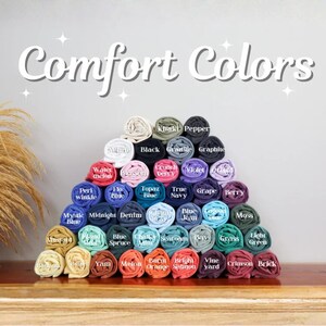 May include: A pyramid of rolled fabric swatches in various colors labeled with text including Ivory, Black, Pepper, Watermelon, Blossom, Crunchberry, Violet, Orchid, Periwinkle, Flo Blue, Topaz Blue, True Navy, Grape, Berry, Mystic Blue, Midnight, Denim, Chambray, Blue Jean, Lagoon Blue, Moss, Mustard, Island Reef, Blue Spruce, Chalky Mint, Seafoam, Bay, Grass, Light Green, Banana, Butter, Yam, Melon, Burnt Orange, Bright Salmon, Vineyard, Crimson, and Brick, with the text Comfort Colors at the top.