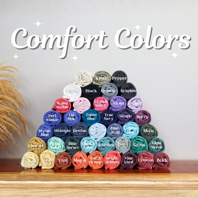 May include: A pyramid of rolled fabric swatches in various colors labeled with text including Ivory, Black, Pepper, Watermelon, Blossom, Crunchberry, Violet, Orchid, Periwinkle, Flo Blue, Topaz Blue, True Navy, Grape, Berry, Mystic Blue, Midnight, Denim, Chambray, Blue Jean, Lagoon Blue, Moss, Mustard, Island Reef, Blue Spruce, Chalky Mint, Seafoam, Bay, Grass, Light Green, Banana, Butter, Yam, Melon, Burnt Orange, Bright Salmon, Vineyard, Crimson, and Brick, with the text Comfort Colors at the top.