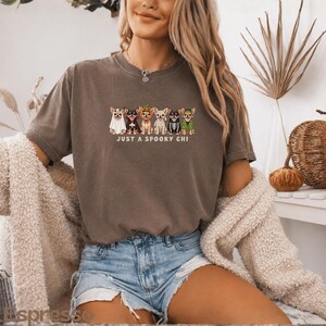 May include: A dark gray short-sleeve t-shirt featuring a graphic of six Chihuahua dogs dressed in various Halloween costumes, including a ghost, vampire, pumpkin, mummy, skeleton, and witch, with the text "JUST A SPOOKY CHI" printed below.