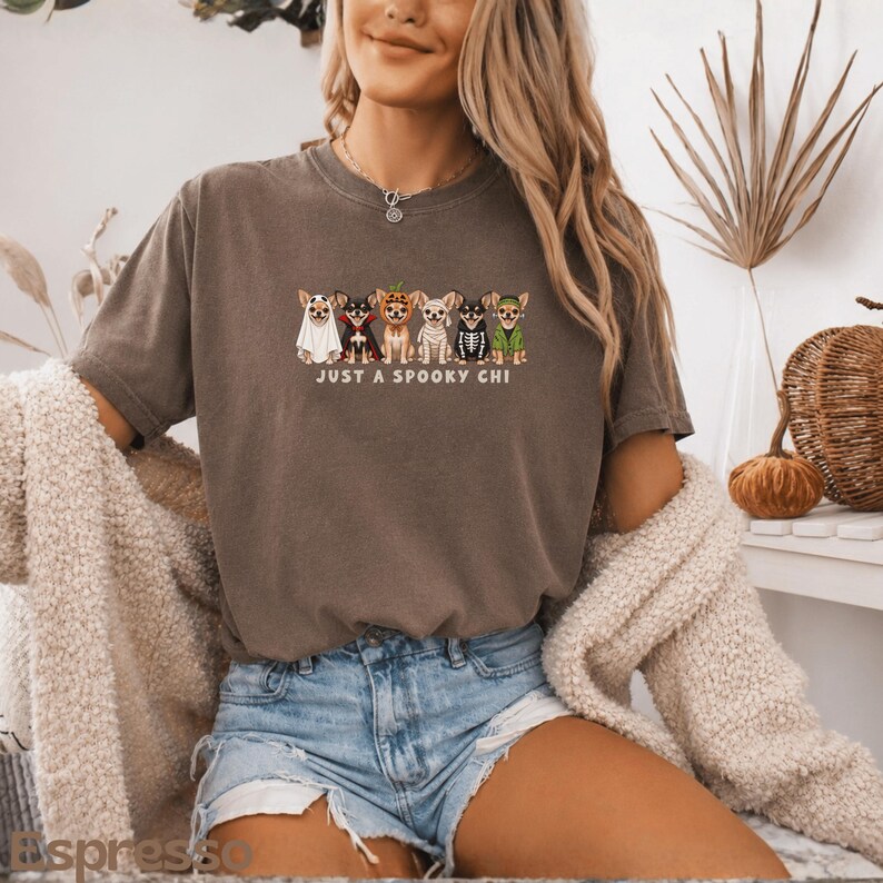 May include: A dark gray short-sleeve t-shirt featuring a graphic of six Chihuahua dogs dressed in various Halloween costumes, including a ghost, vampire, pumpkin, mummy, skeleton, and witch, with the text "JUST A SPOOKY CHI" printed below.