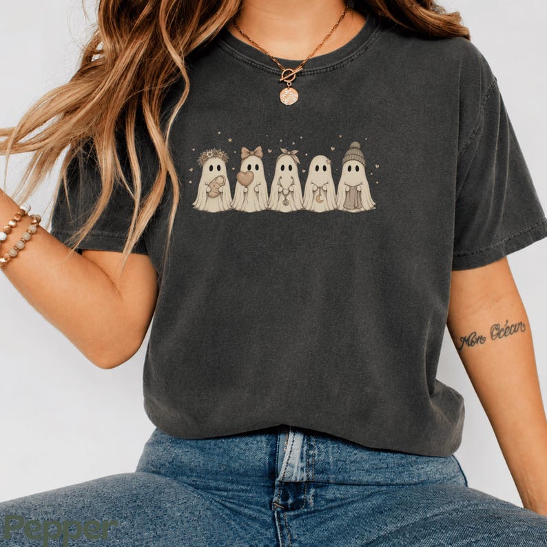 May include: A charcoal grey short-sleeve t-shirt featuring a graphic print of five ghosts in a row. Each ghost has a different accessory, including a floral crown, a bow, a headband, a crescent moon necklace, and a knit beanie hat.