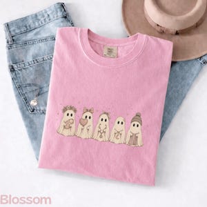 May include: A light pink t-shirt featuring a graphic of five beige ghosts in a row. The ghosts are decorated with a flower crown, a bow, a headband, a crescent moon charm, and a knit beanie. The shirt has a crew neckline and a tag at the collar.