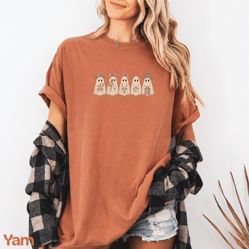 May include: A rust-coloured short-sleeve t-shirt featuring a graphic of five ghosts in a row, each with different accessories including a flower crown, bow, necklace, and beanie. The shirt is worn with a black and white plaid flannel shirt tied around the waist.