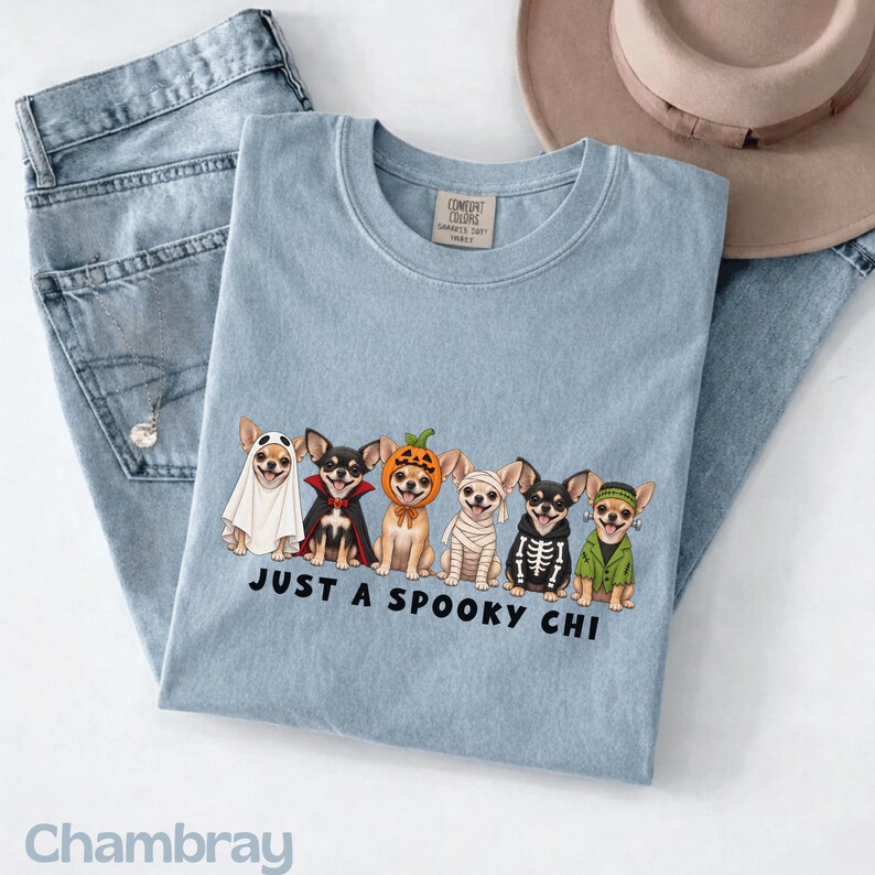 May include: A light blue t-shirt featuring a graphic of six Chihuahua dogs dressed in various Halloween costumes, including a ghost, vampire, pumpkin, mummy, skeleton, and Frankenstein. The text below the dogs reads, "JUST A SPOOKY CHI."