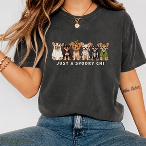 May include: A charcoal gray short-sleeve t-shirt featuring a graphic of six Chihuahua dogs in various Halloween costumes, including a ghost, vampire, pumpkin, mummy, skeleton, and green shirt. The text below the dogs reads JUST A SPOOKY CHI.