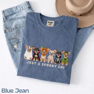 May include: A blue t-shirt featuring a graphic of six Chihuahua dogs dressed in various Halloween costumes, including a ghost, vampire, pumpkin, mummy, skeleton, and Frankenstein's monster, with the text "JUST A SPOOKY CHI" printed below.