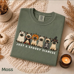 Op de afbeelding: Een mosgroen T-shirt met ronde hals en een afbeelding van zes Siamezen in verschillende Halloween-kostuums, waaronder een geest, vampier, pompoen, mummie, skelet en heks. De tekst onder de katten luidt: "JUST A SPOOKY SIAMESE."