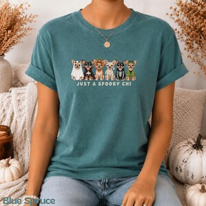 May include: A teal short-sleeve t-shirt featuring a graphic of six Chihuahua dogs dressed in various Halloween costumes, including a ghost, vampire, pumpkin, mummy, skeleton, and witch. The text below the dogs reads JUST A SPOOKY CHI.