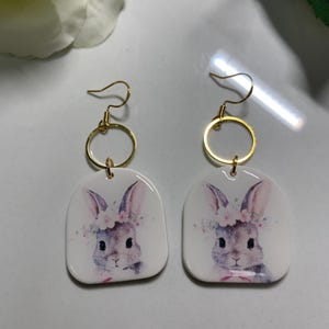 May include: A pair of white bunny earrings with gold hoops and hooks. The bunnies have pink noses and are wearing flower crowns.