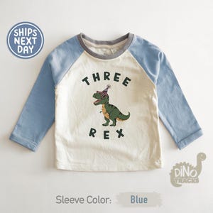 Pu&ograve; includere: Una maglietta a maniche lunghe blu chiaro e color crema con una grafica di dinosauro. La maglietta presenta la scritta "THREE REX" con un dinosauro dei cartoni animati con occhiali da sole e un cappello da festa. Le maniche sono blu chiaro.