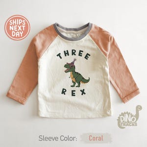 Pu&ograve; includere: Maglietta a maniche lunghe bianco sporco e color corallo con la scritta "THREE REX" e un dinosauro dei cartoni animati con occhiali da sole e un cappello da festa. La maglietta ha uno scollo grigio e maniche color corallo.