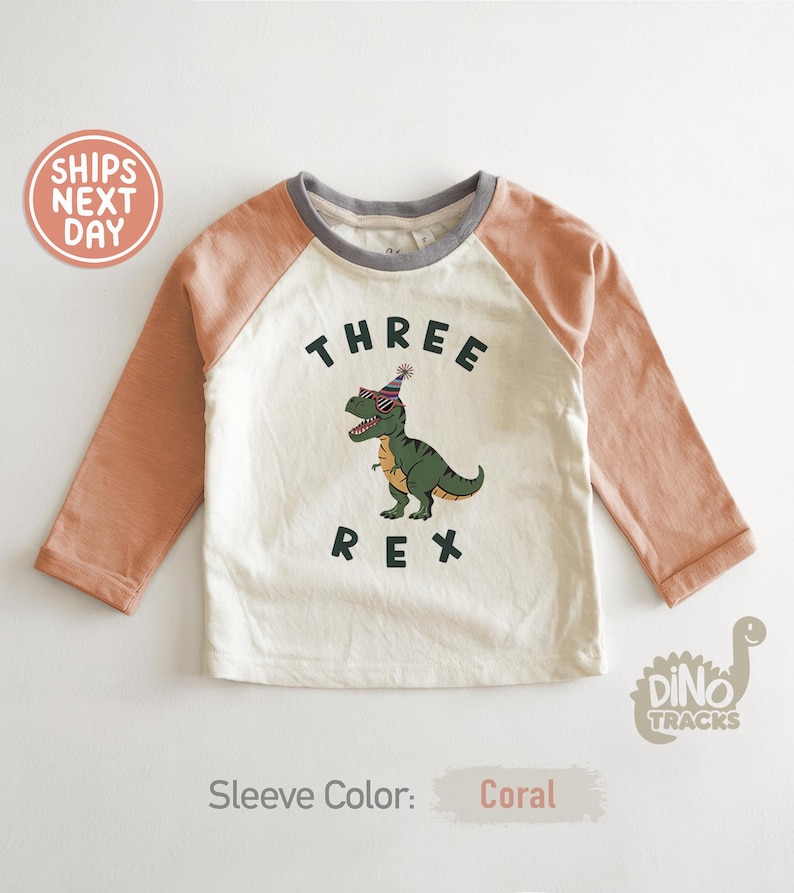 Pu&ograve; includere: Maglietta a maniche lunghe bianco sporco e color corallo con la scritta "THREE REX" e un dinosauro dei cartoni animati con occhiali da sole e un cappello da festa. La maglietta ha uno scollo grigio e maniche color corallo.