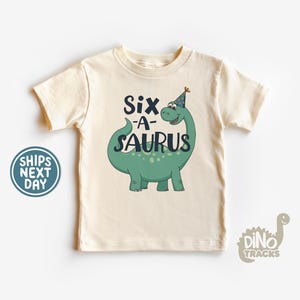 May include: Cream-colored t-shirt with a green dinosaur graphic and the text "Six-A-Saurus." The dinosaur wears a party hat. A round blue and white sticker says "Ships Next Day." The Dino Tracks logo is in the bottom right corner.