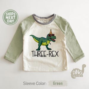 May include: A long-sleeved, raglan-style shirt with a cream-colored body and green sleeves. The shirt features a graphic of a green dinosaur wearing sunglasses and a party hat, with the text "THREE-REX" below. The sleeve color is green.
