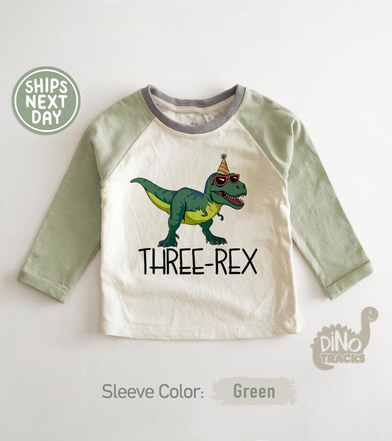 May include: A long-sleeved, raglan-style shirt with a cream-colored body and green sleeves. The shirt features a graphic of a green dinosaur wearing sunglasses and a party hat, with the text "THREE-REX" below. The sleeve color is green.
