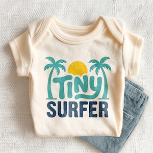 May include: A cream-colored short-sleeve baby bodysuit featuring a graphic print of two teal palm trees, a yellow sun, and the text "TINY SURFER" in teal and dark blue. The garment is paired with a piece of blue corduroy fabric visible at the side.
