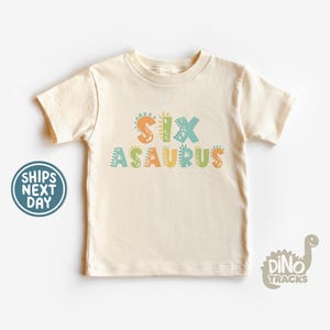 May include: A light beige t-shirt with the text "SIXASAURUS" in colorful letters with dinosaur footprints. The letters are in shades of green, blue, orange, and yellow.