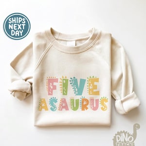 May include: A beige sweatshirt with the text "FIVE-ASAURUS" in colorful dinosaur-shaped letters. The letters are outlined in black and have a speckled pattern.