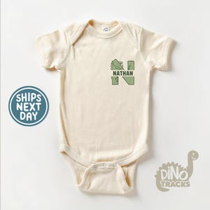May include: A cream-colored baby onesie with a green dinosaur graphic forming the letter N and the name "NATHAN" printed below. The onesie has a snap closure at the bottom. The image also includes a logo with the text "Dino Tracks" and a cartoon dinosaur.