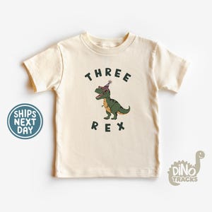 Pu&ograve; includere: T-shirt color crema per bambini piccoli con una grafica di un dinosauro verde con occhiali da sole e un cappello da festa. Il testo "THREE REX" &egrave; stampato sopra e sotto il dinosauro.