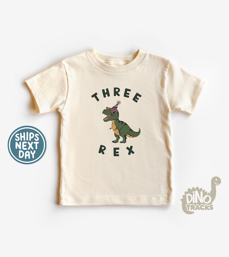 Pu&ograve; includere: T-shirt color crema per bambini piccoli con una grafica di un dinosauro verde con occhiali da sole e un cappello da festa. Il testo "THREE REX" &egrave; stampato sopra e sotto il dinosauro.