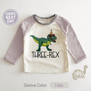 May include: A white and lilac long-sleeve shirt featuring a green cartoon Tyrannosaurus Rex wearing sunglasses and a party hat. The text "THREE-REX" is printed below the dinosaur. The sleeve color is lilac.
