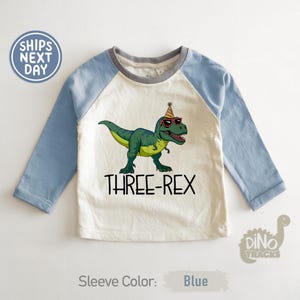 May include: A white and blue raglan-style shirt with a green dinosaur graphic wearing sunglasses and a party hat. The text "THREE-REX" is below the dinosaur. The sleeve color is blue.