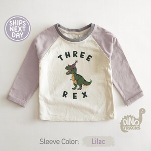 Pu&ograve; includere: Una maglietta a maniche lunghe in stile raglan con corpo color crema e maniche lilla. La maglietta presenta una grafica di un dinosauro verde con occhiali da sole e un cappello da festa, con le parole "THREE REX" stampate.