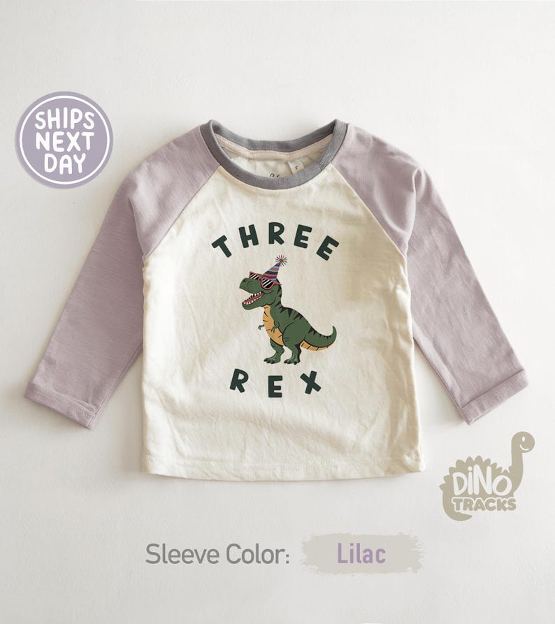 Pu&ograve; includere: Una maglietta a maniche lunghe in stile raglan con corpo color crema e maniche lilla. La maglietta presenta una grafica di un dinosauro verde con occhiali da sole e un cappello da festa, con le parole "THREE REX" stampate.