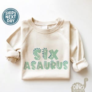 May include: A cream-colored sweatshirt with the text "SIXASAURUS" in green letters with dinosaur-shaped outlines. The letters are outlined in black.