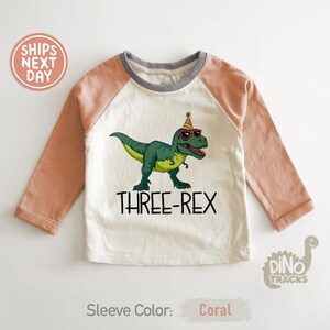 May include: A long-sleeved, coral and cream-colored shirt with a cartoon dinosaur wearing sunglasses and a party hat. The text "THREE-REX" is below the dinosaur. The shirt has a gray neckline.