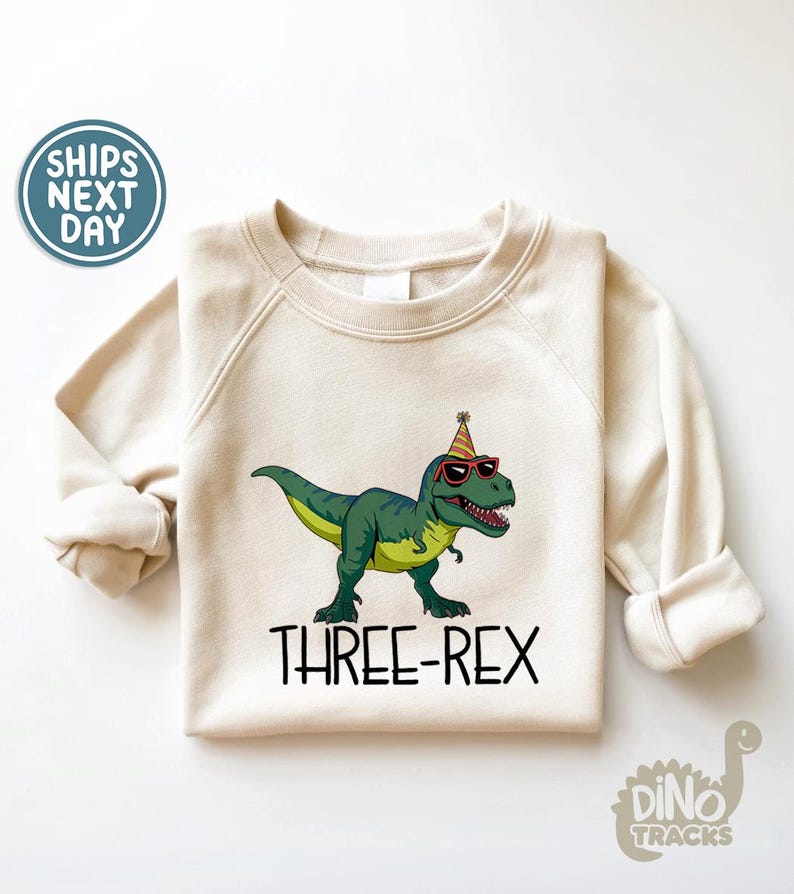 May include: Cream-colored sweatshirt featuring a green dinosaur wearing sunglasses and a party hat. The text "THREE-REX" is printed below the dinosaur. The sweatshirt has long sleeves and a crew neck.