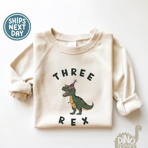 Pu&ograve; includere: Felpa color crema con la scritta "THREE REX" in nero. Un dinosauro dei cartoni animati con occhiali da sole e un cappello da festa &egrave; al centro. La felpa ha maniche lunghe e girocollo.