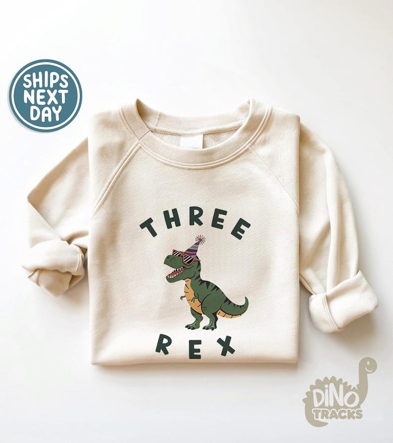 Pu&ograve; includere: Felpa color crema con la scritta "THREE REX" in nero. Un dinosauro dei cartoni animati con occhiali da sole e un cappello da festa &egrave; al centro. La felpa ha maniche lunghe e girocollo.