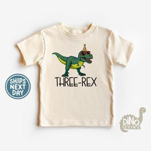 May include: Cream-colored t-shirt featuring a green dinosaur wearing sunglasses and a party hat. The text "THREE-REX" is printed in black below the dinosaur. A fun birthday-themed graphic tee.