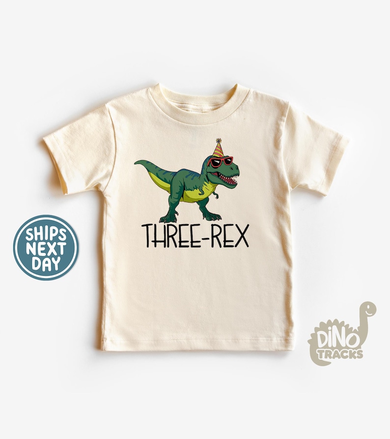 May include: Cream-colored t-shirt featuring a green dinosaur wearing sunglasses and a party hat. The text "THREE-REX" is printed in black below the dinosaur. A fun birthday-themed graphic tee.