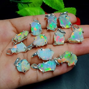 Puede incluir: Una colección de colgantes de ópalo con engastes plateados. Los ópalos muestran una gama de colores, incluyendo verde, naranja y rosa, sobre una base blanca. Cada colgante es único en forma y tamaño, mostrando la belleza natural de las piedras preciosas.