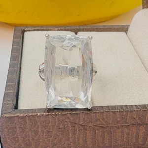 May include: A clear, rectangular-cut gemstone ring. The large, faceted stone is set on a silver band. The ring is displayed in a brown jewelry box with a cream-colored interior. The gemstone is approximately 1.5 inches in length.