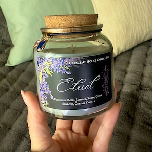 Może przedstawiać: Świeca w szklanym słoiku z korkową pokrywką i ozdobnym urokiem. Etykieta głosi "Etriel" z nutami zapachowymi Zakazaną Różą, Jaśminem, Cukrem, Cedrem, Cieniami i Kremową Wanilią. Świeca jest trzymana w dłoni.