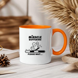 May include: White ceramic mug with an orange rim. The mug has a black graphic of a butterfly with the text "Miracle Buffering" and "Please Wait..." below the butterfly.