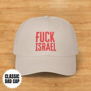 Puede incluir: Gorra de béisbol clásica beige claro con la inscripción roja "FUCK ISRAEL". También es visible una pegatina blanca con "CLASSIC DAD CAP". Fondo de madera.