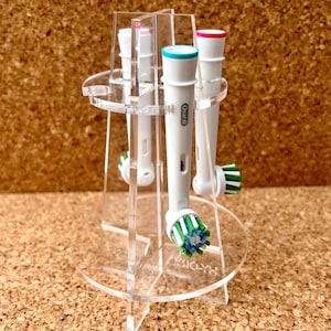 May include: A clear acrylic toothbrush holder with four toothbrush heads. The toothbrush heads are white with green and blue bristles. The holder has a round base and three legs.