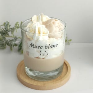 Peut inclure: Une bougie en verre avec un design en couches. La base est marron clair, surmontée de cire fouettée blanche et d'une boule de cire rose pâle. La bougie porte le texte "Muse blanc" et repose sur un petit dessous de verre en bois.