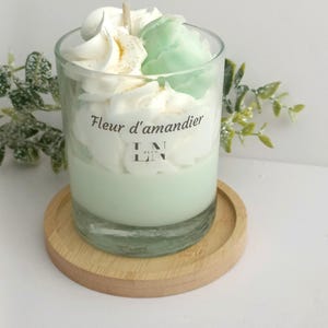 Peut inclure: Une bougie en verre transparent avec une base vert menthe et une garniture en cire ressemblant à de la crème fouettée. La bougie porte l'inscription "Fleur d'amandier" sur le verre et repose sur un petit dessous de verre en bois.