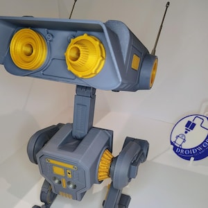 Könnte beinhalten: Ein grau-gelber 3D-gedruckter Roboter mit Antennen. Der Roboter hat zwei große gelbe Augen und einen gelben Streifen auf der Brust. Der Roboter steht auf einer weißen Oberfläche.