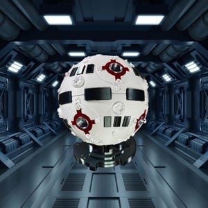 May include: A white spherical object featuring black rectangular panels and red circular details. The object has a textured surface and appears to be floating. The background is a futuristic, metallic corridor with blue lighting.