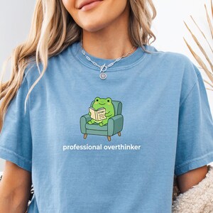 May include: A light blue t-shirt with the words "professional overthinker" printed on it. The shirt features a cartoon frog reading a book while sitting in a small armchair. The shirt is made of a soft material.