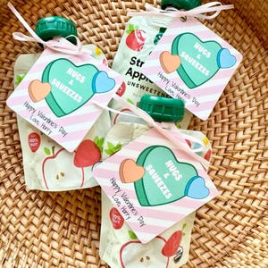 May include: Three pink and white Valentine's Day gift tags with the text "Hugs & Squeezes" and a heart-shaped candy design. Each tag is tied to a pouch of apple sauce with a green lid.