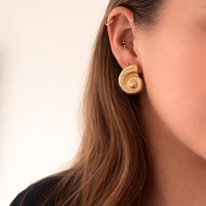 Peut inclure: Boucles d'oreilles dorées en forme de spirale. Les boucles d'oreilles présentent un motif texturé et détaillé, ressemblant à une coquille de nautile. Les boucles d'oreilles sont portées sur l'oreille, avec la tige visible. Le fond est de couleur neutre.