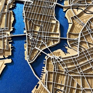 Puede incluir: Un mapa de madera detallado y en capas de la ciudad de Nueva York, que muestra la disposición de la cuadrícula de la ciudad y las vías fluviales. El mapa está hecho de madera de color claro con líneas blancas que representan las calles, sobre un fondo azul. Se ven puentes y el contorno de la ciudad.